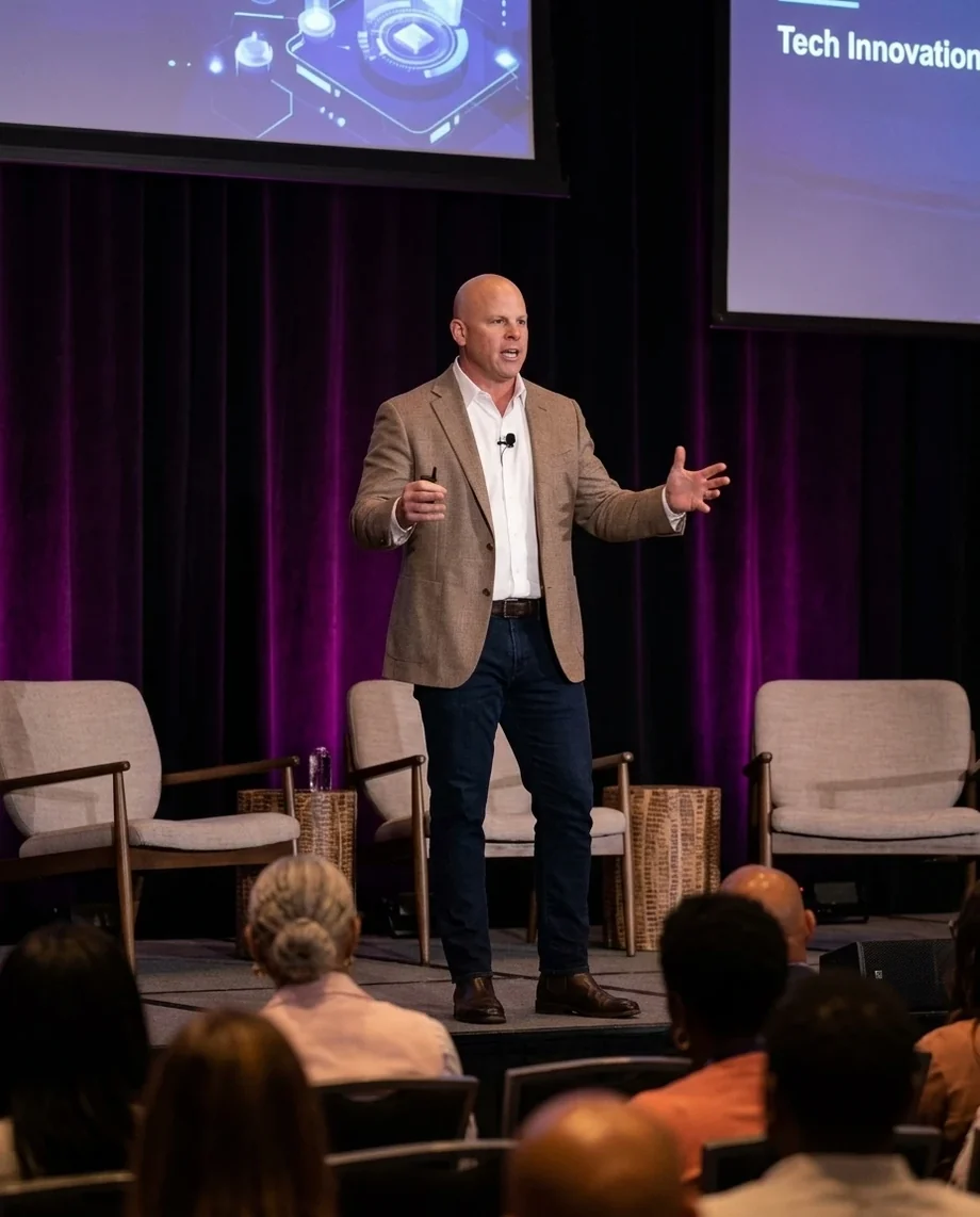 Damon Wells speaking on a stage in front of a seated audience, with armchairs set for a panel behind him.